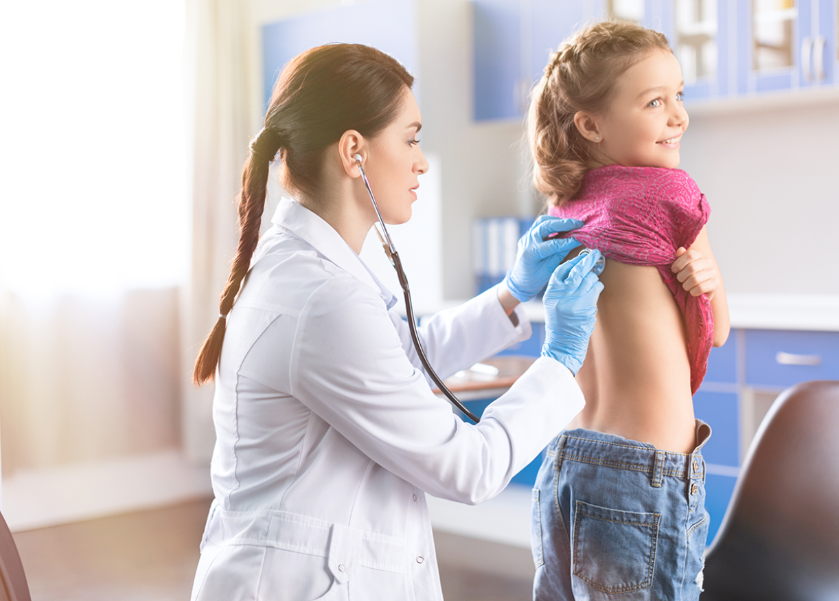 A doctor using a stethoscope to examine a smiling child's back in a medical offi