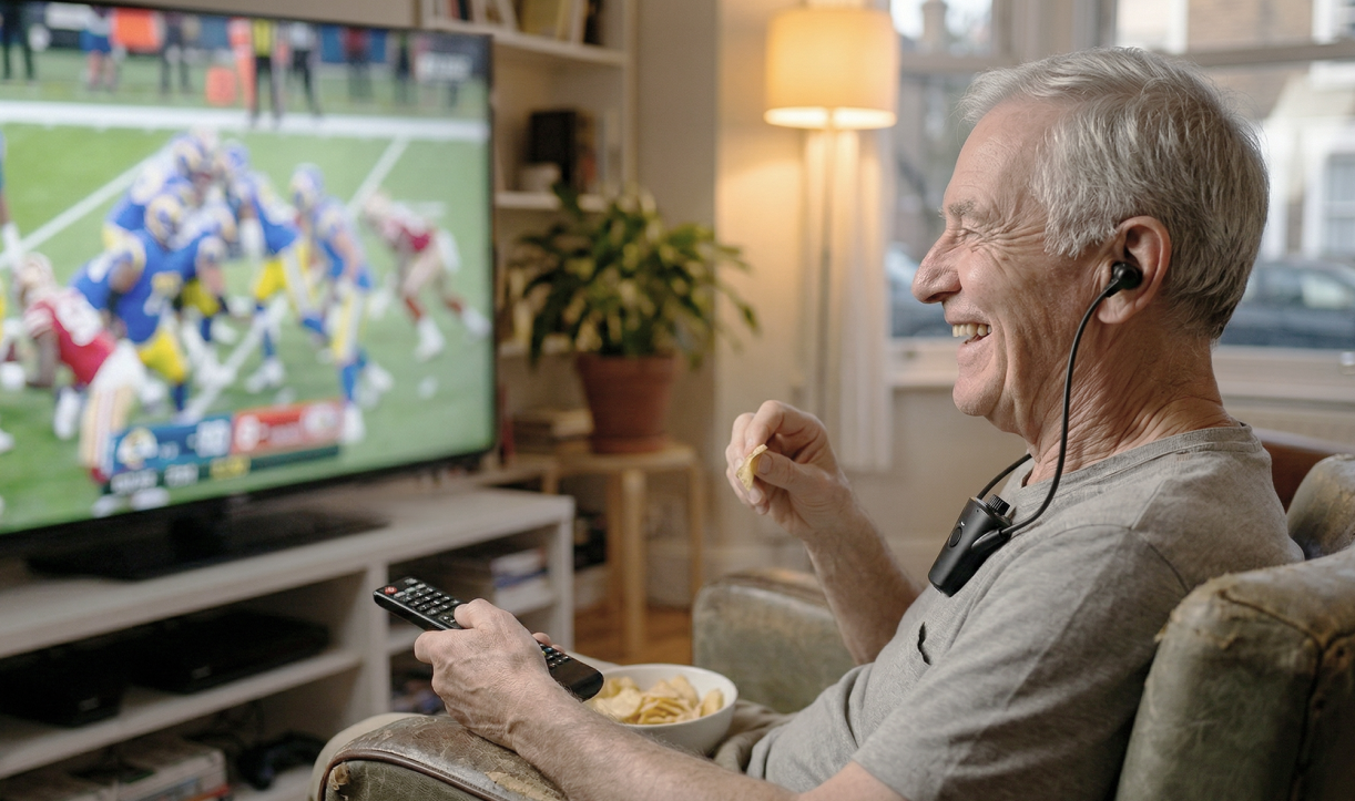 Older man using HearDirect headset while watching a football game and eating snacks