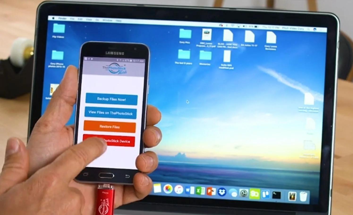 Hand holding a phone with ThePhotoStick Mobile app open in front of a laptop screen